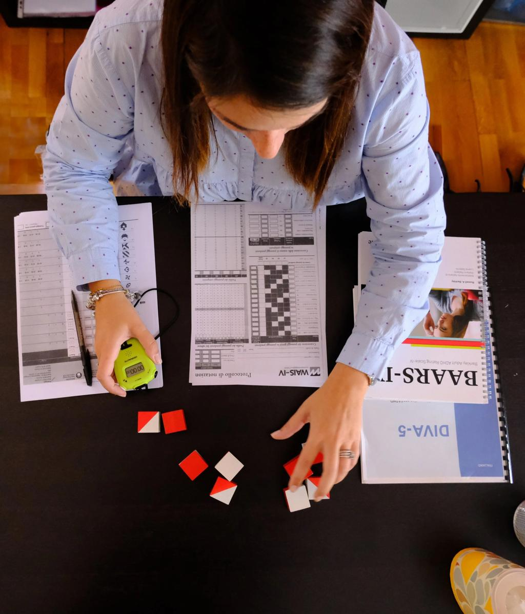 Vista superior de uma mesa durante uma avaliação neuropsicológica, com protocolos de registro, cronômetro e blocos de teste sendo manipulados
